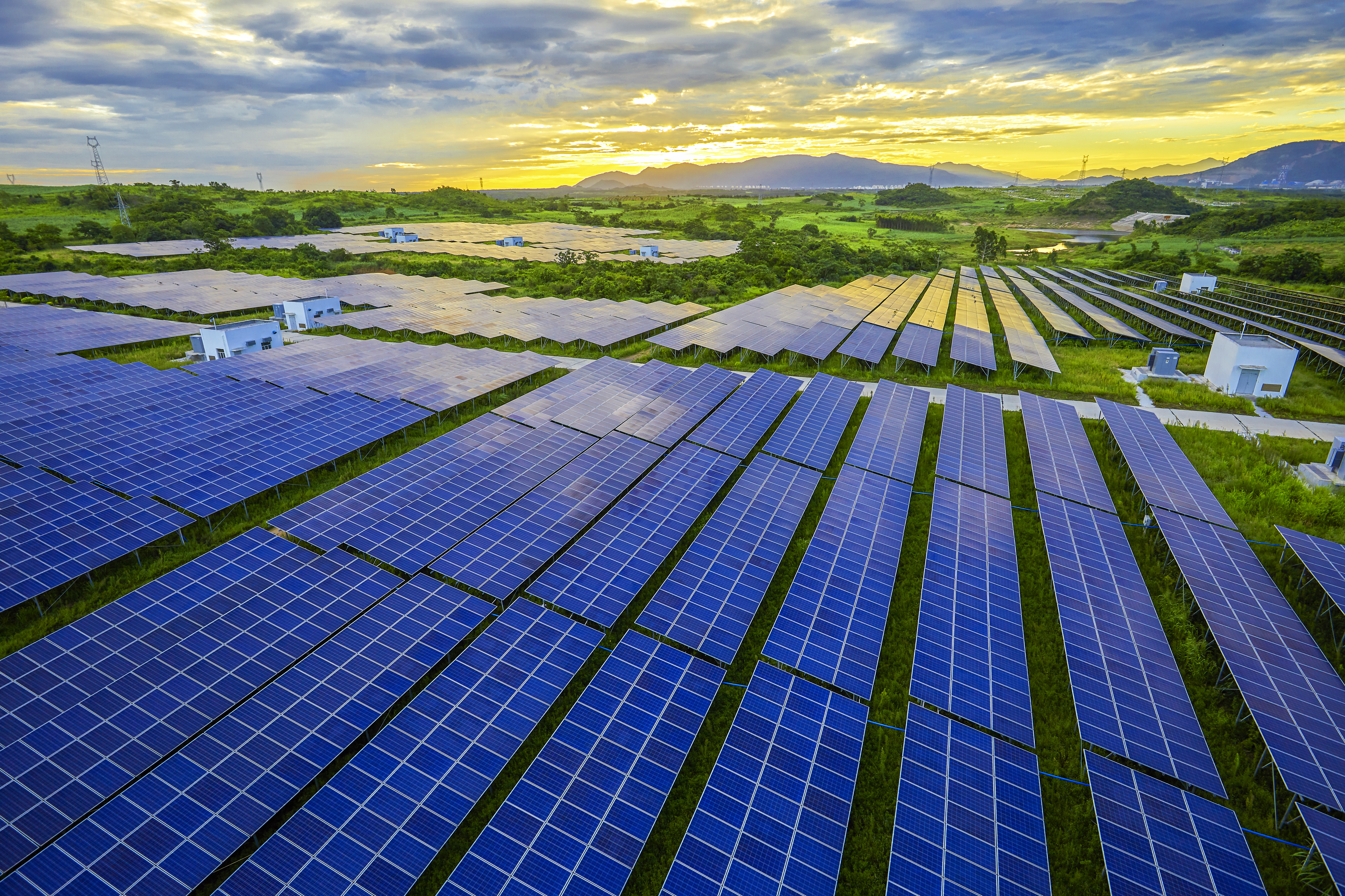 Solar panels in a large photovoltaic field capturing sunlight during sunset, with green fields and distant mountains under a partly cloudy sky.