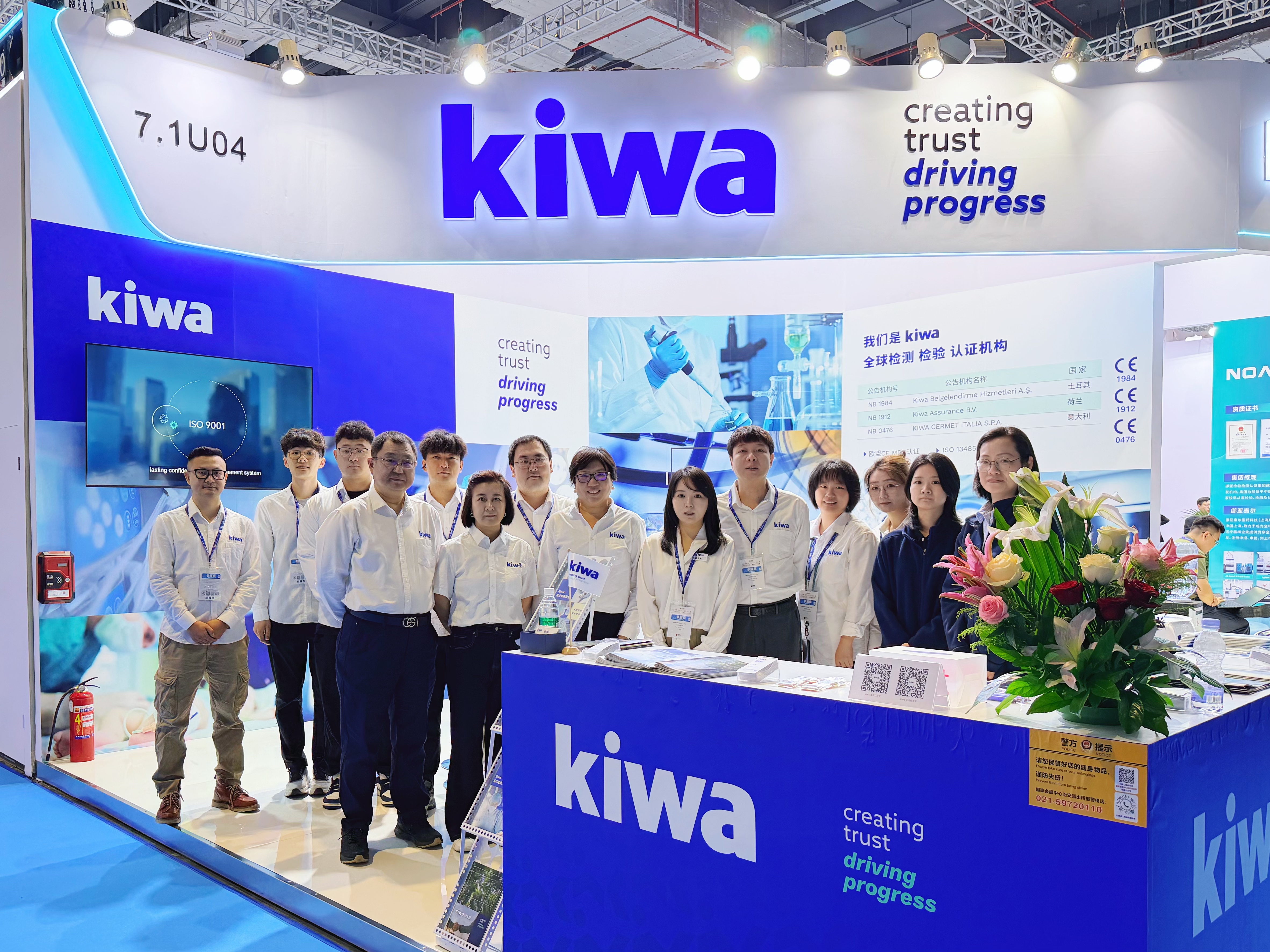 Kiwa展台前的一群工作人员合影，背景为蓝白色企业展位和“creating trust driving progress”标语，前台摆放花束和宣传资料。