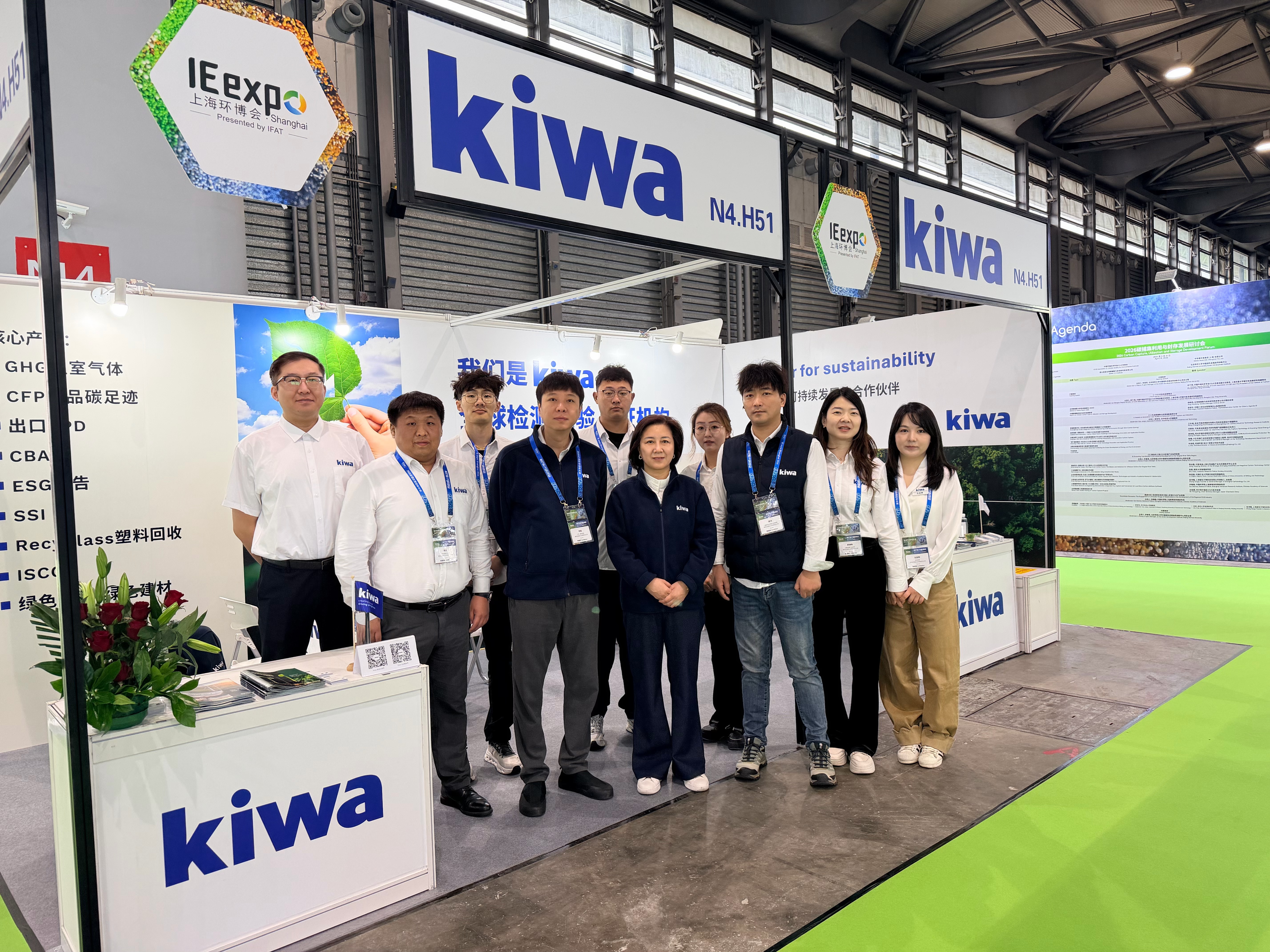 上海国际环保展览会上的 Kiwa 展台，多名工作人员身穿工牌合影，背景展示 IEexpo Shanghai 和 sustainability 相关宣传板。
