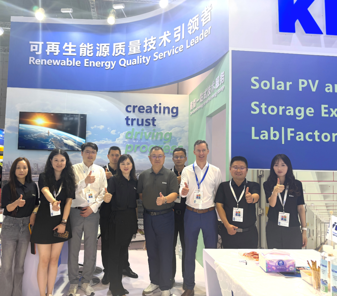 展会展台前的一组Kiwa同事合影，背景展示“Renewable Energy Quality Service Leader”“Solar PV and Storage”等太阳能与储能相关宣传标语。多人佩戴证件并竖起大拇指，桌上摆放宣传资料和样品。