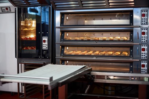 Commercial bakery oven with multiple trays of bread rolls baking inside, alongside a separate heating unit and a metal worktable.