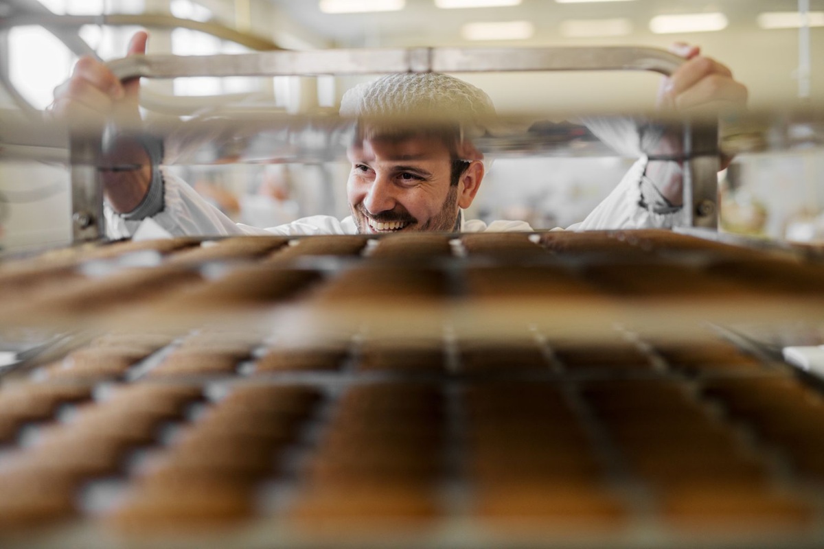 Person in a white coat handling trays of baked goods in a bakery or food production setting.