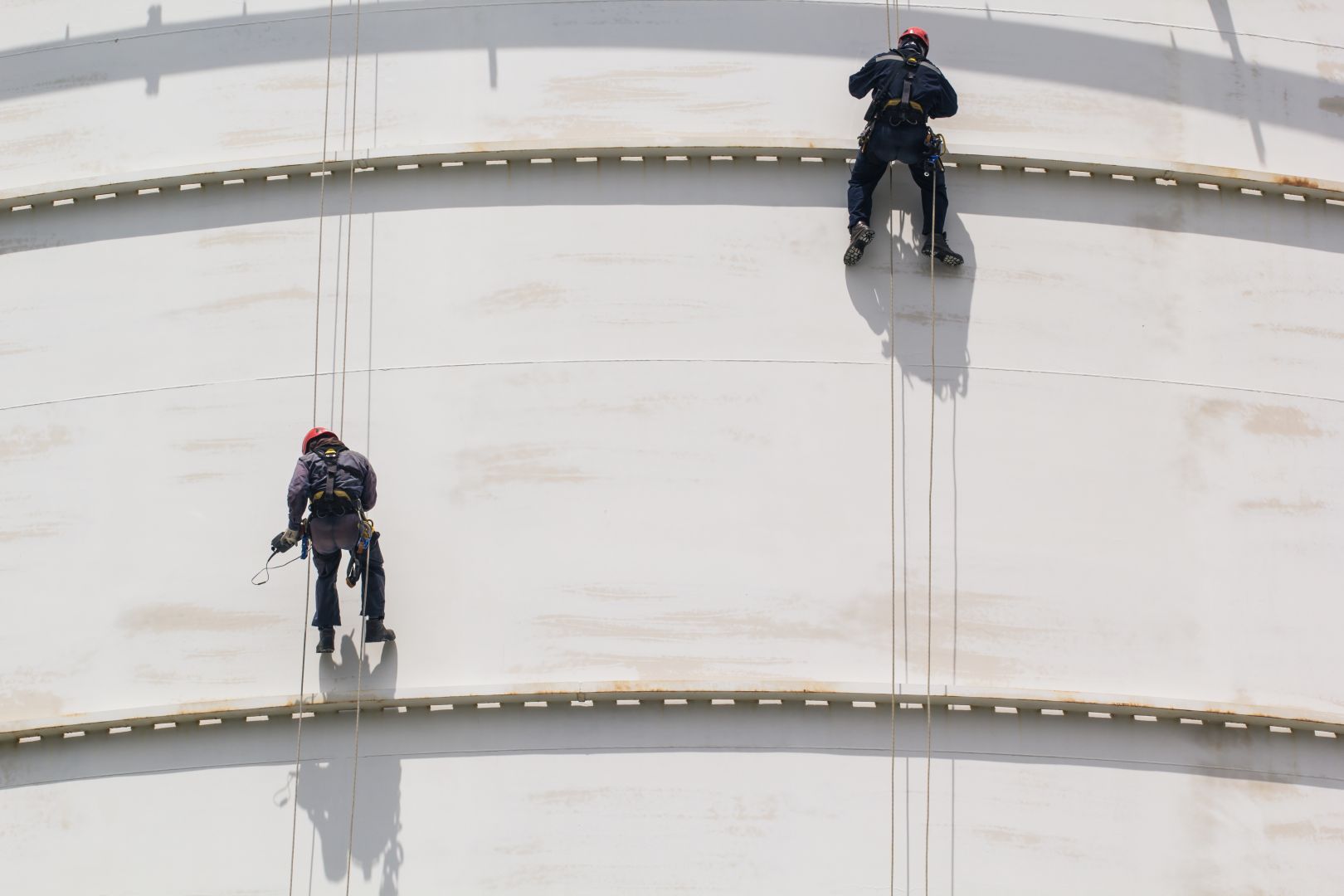 Twee industriële klimmers met veiligheidsuitrusting hangen aan touwen tegen een grote witte metalen tank.