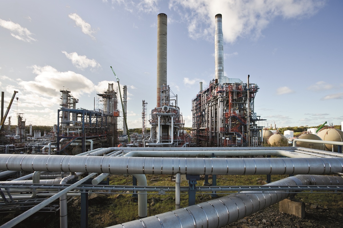 Industrieel energiecomplex met grote pijpleidingen, metalen installaties en twee hoge schoorstenen onder een deels bewolkte hemel.