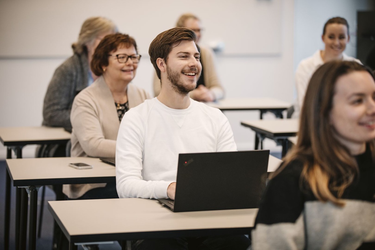 Groep volwassenen in een klaslokaal met laptops aan tafels tijdens een training of vergadering.