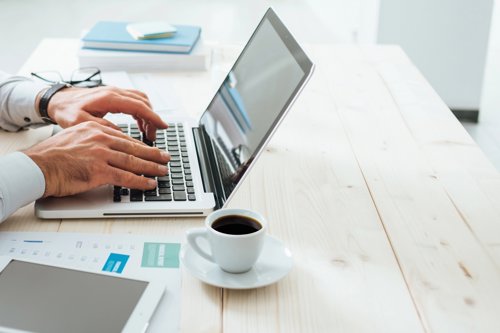 Man typing on a laptop at a wooden desk with coffee cup, tablet, and documents nearby.