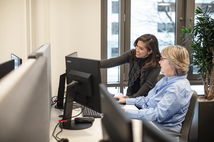 Two office workers reviewing a computer monitor in a modern office workspace with desks, monitors, and a potted plant.
