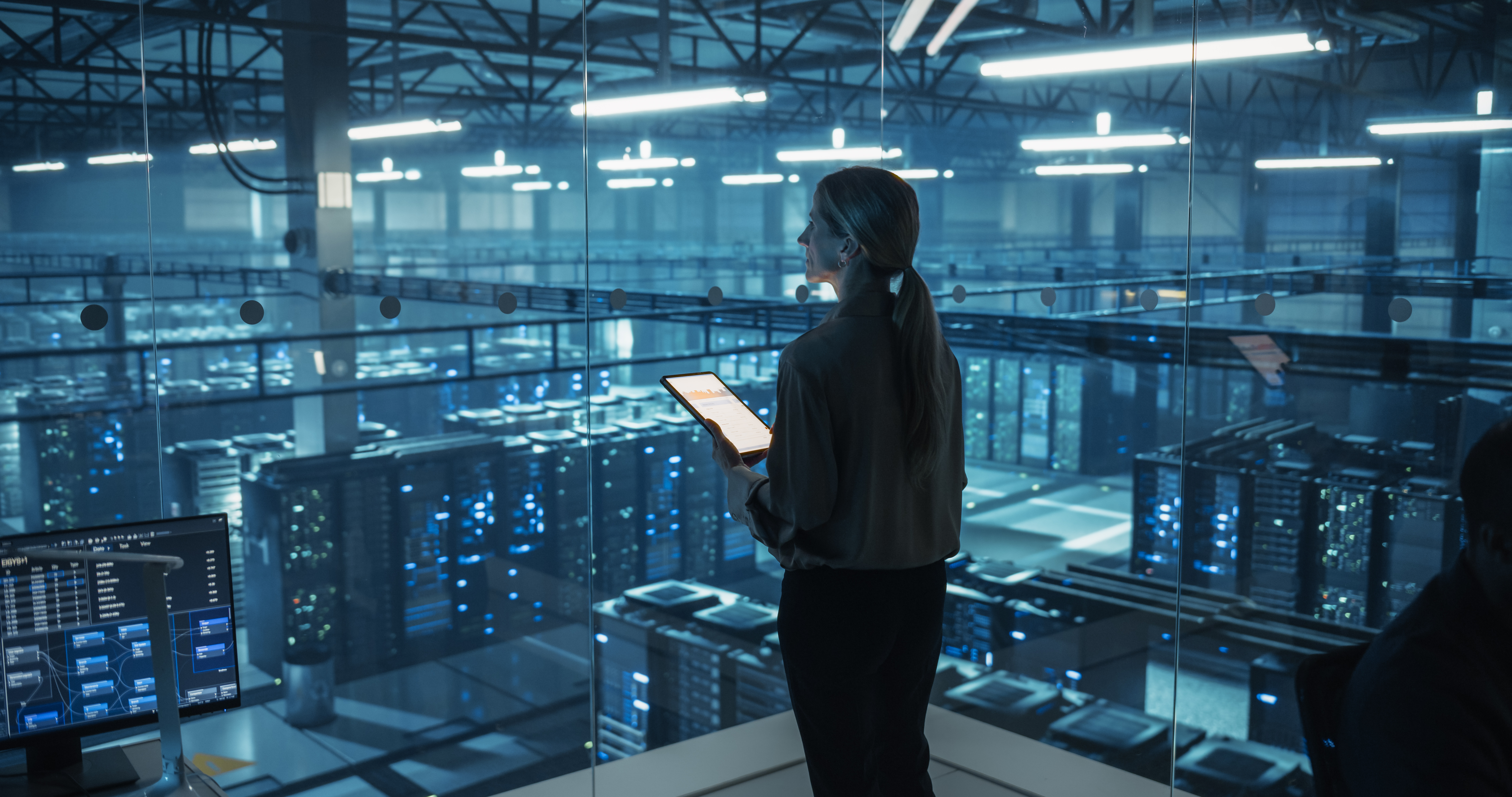 Person med surfplatta som står och tittar ut över ett modernt datacenter med serverrack och blå belysning, i en bild som förmedlar IT och AI inom Affärssäkerhet.