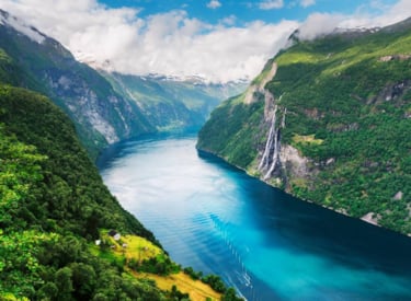 Naturbild visar en fjord mellan gröna höga berg som har snötäckta toppar längst bort i horisonten.