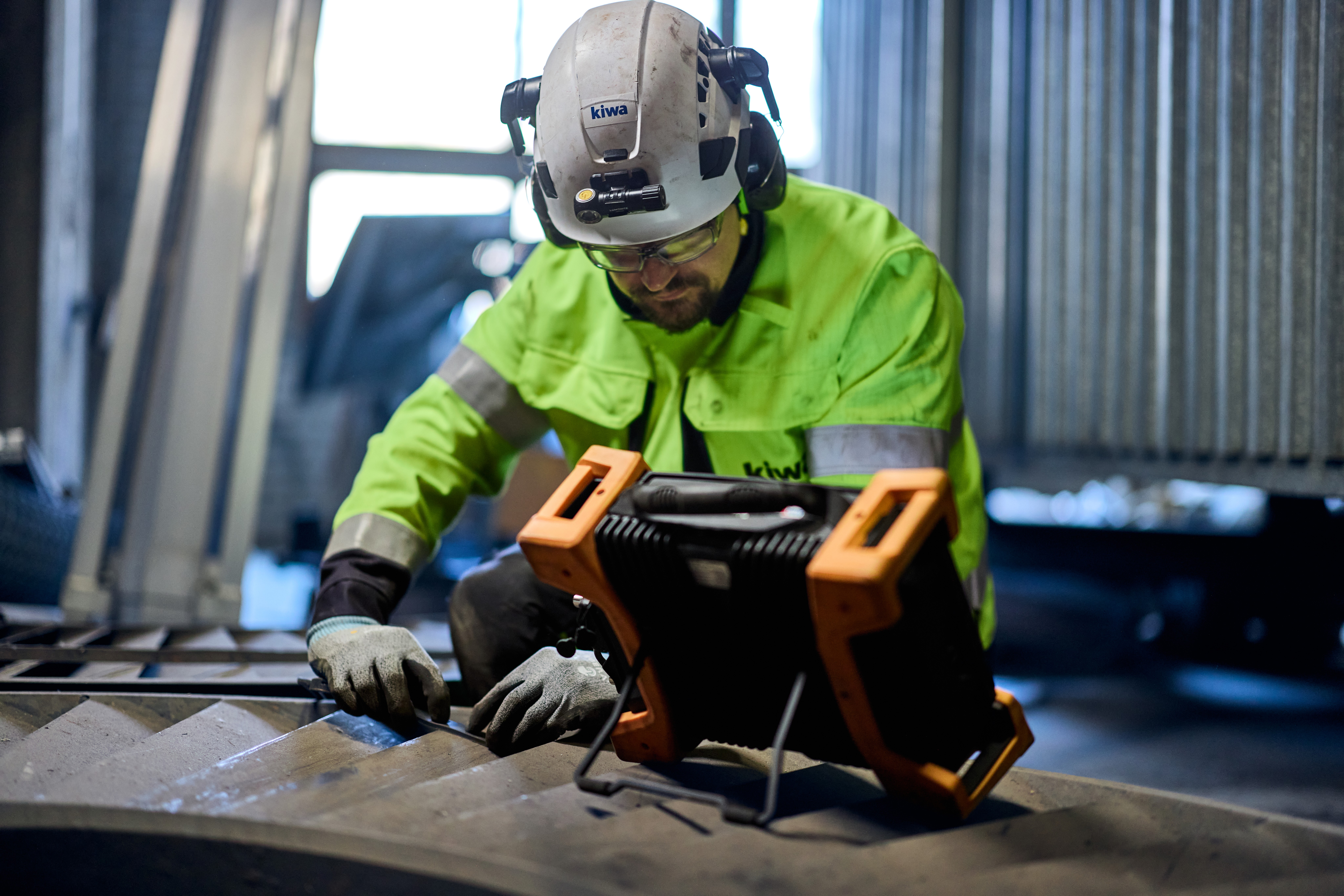 Arbetare i reflexjacka och hjälm inspekterar en metallkonstruktion i industriell miljö.