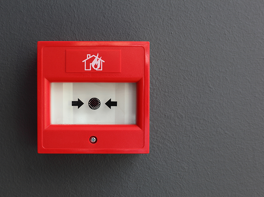 Rød brandalarm på en grå væg med pile og brandsymbol.