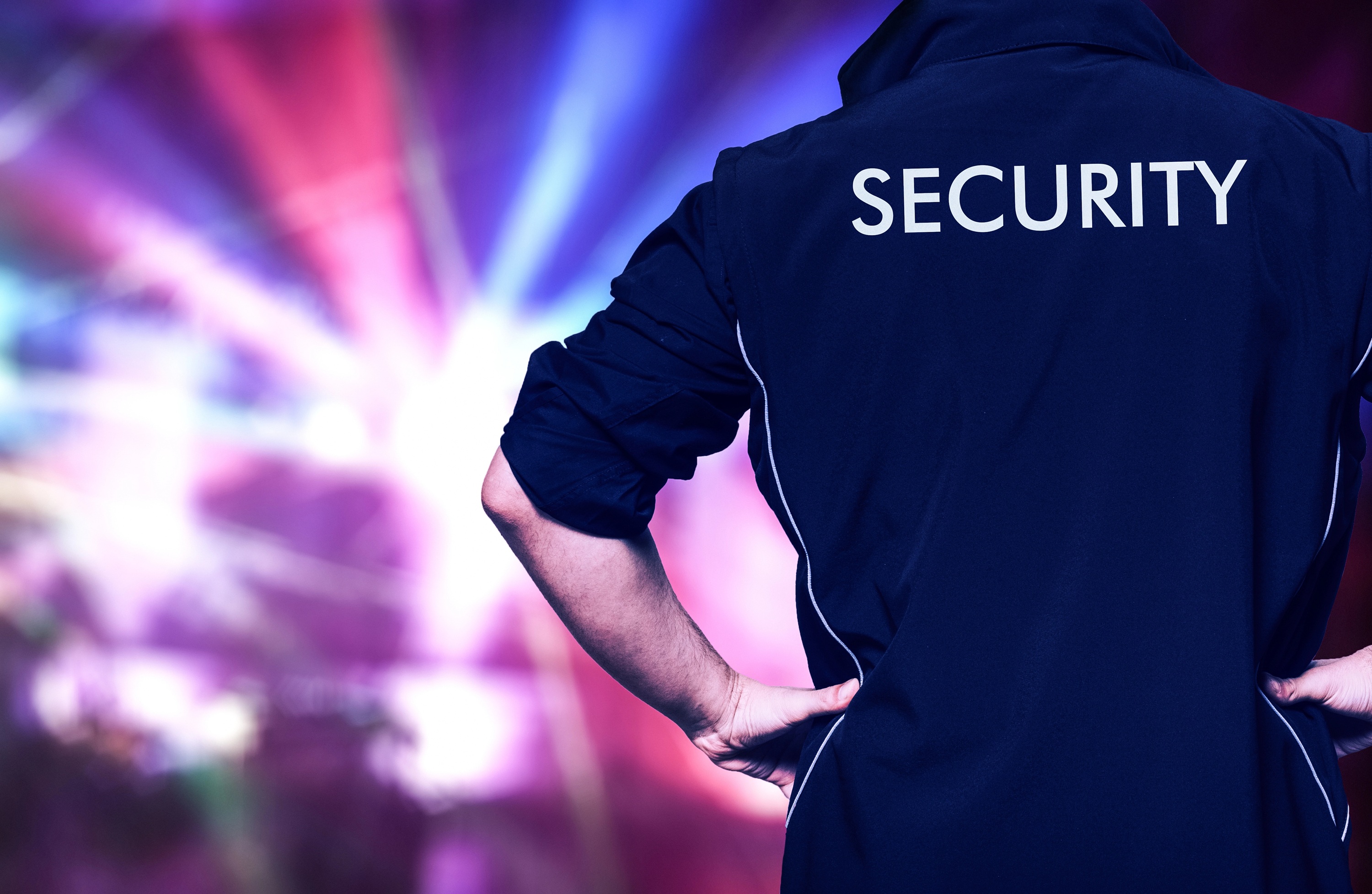 Man in een donkerblauw jack met de tekst "SECURITY" op de rug, staande voor een kleurrijke achtergrond met lichteffecten.