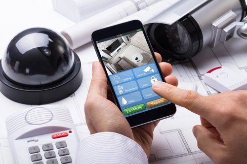 Persoon die een smartphone gebruikt om een beveiligingssysteem te bedienen, omringd door beveiligingsapparatuur zoals camera's en een alarmsysteem, met technische tekeningen op de achtergrond.
