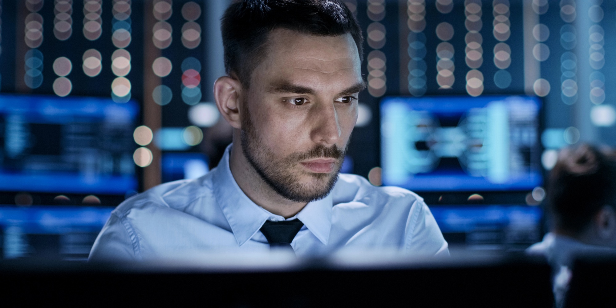 Man zittend aan een bureau in een moderne controlekamer, met achtergrond van computerschermen en LED-verlichting.