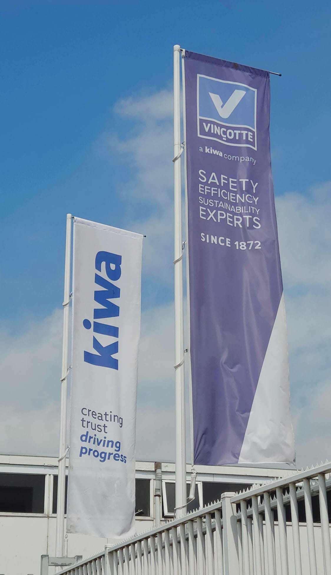 Twee wapperende vlaggen van Kiwa en Vinçotte tegen een blauwe lucht, met de teksten ‘creating trust driving progress’ en ‘Safety Efficiency Sustainability Experts’.