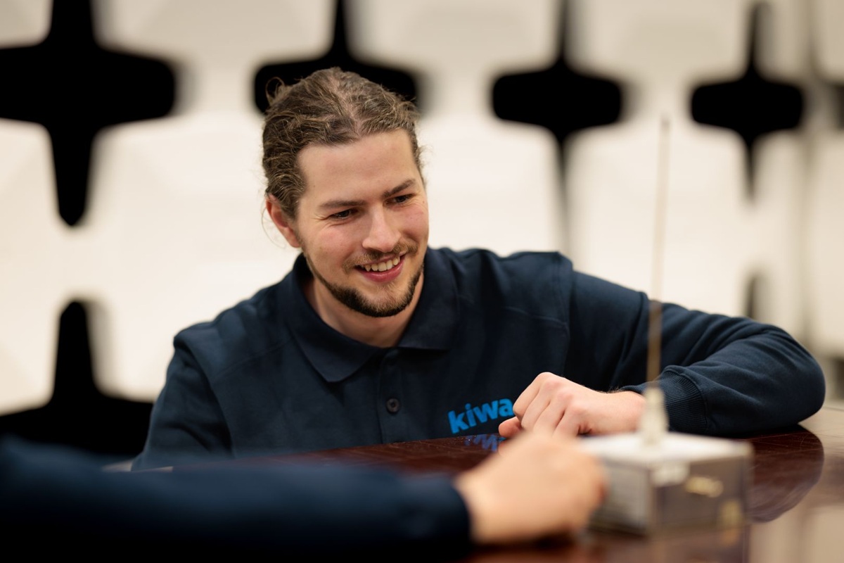 Portrait of a person in a dark blue Kiwa polo shirt seated at a table in an office setting.