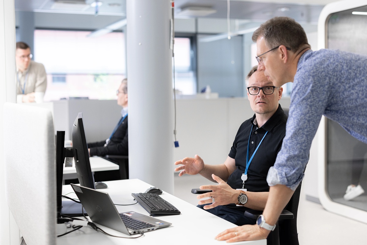 Twee mannen overleggen bij een bureau met laptop en monitor in een modern kantoor.