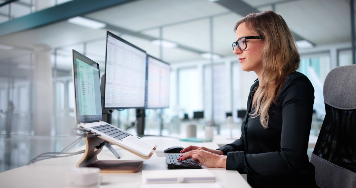 Vrouw op kantoor achter een bureau met meerdere computerschermen en een toetsenbord, werkend in een moderne kantooromgeving.