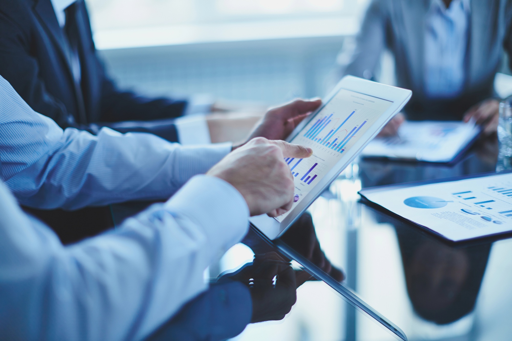 Business meeting with people reviewing graphs and charts on a tablet and documents at a glass table.