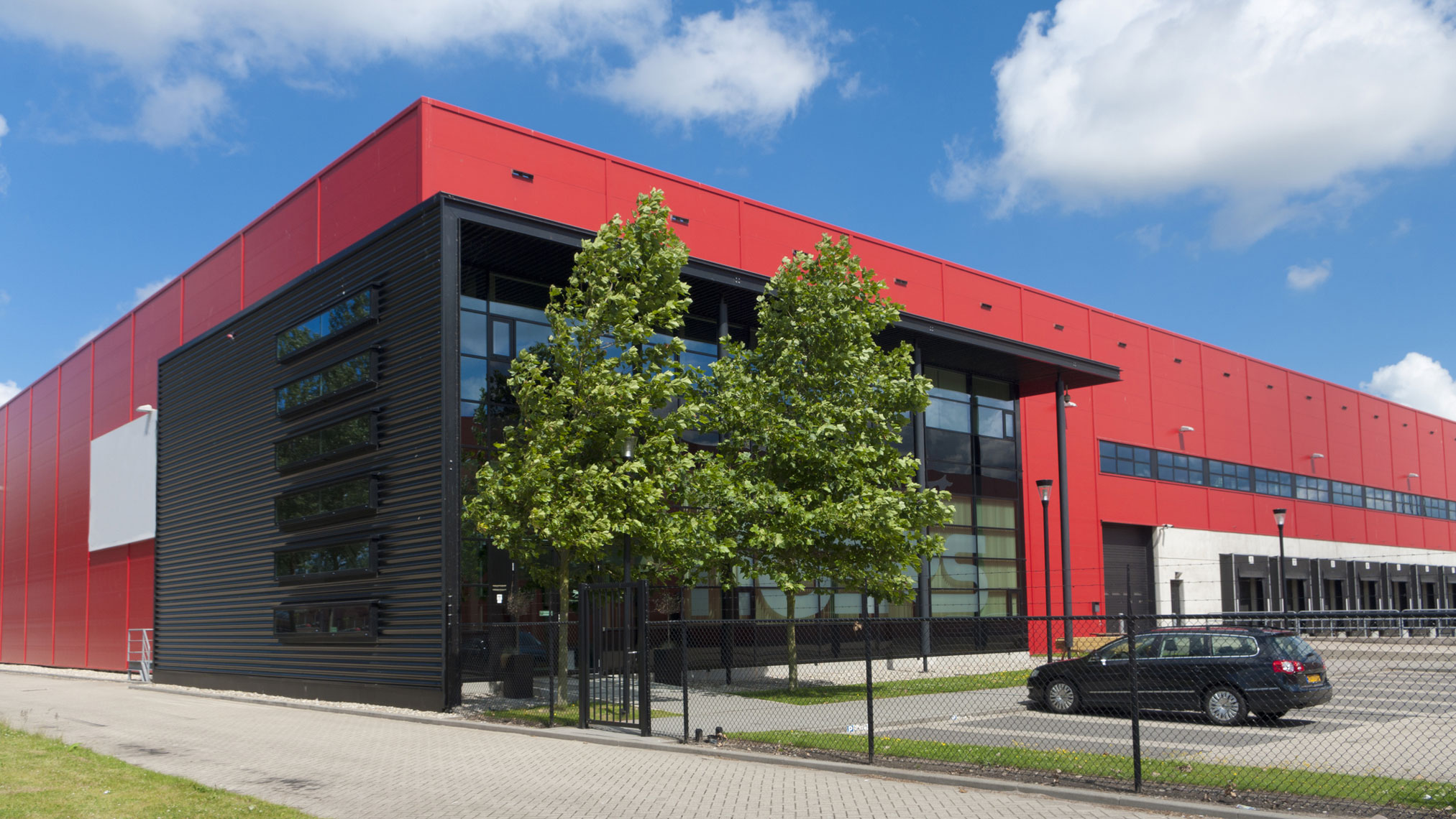 Stor röd och svart industribyggnad med stora fönster, träd framför entrén.