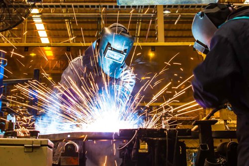Två svetsare med svetshjälm arbetar i en industrimiljö med kraftiga gnistor under svetsning - med hjälp av Kiwas tjänst för svetsvalidering enligt IEC 60974-14 får du både validering och kalibrering av din svetsutrustning.