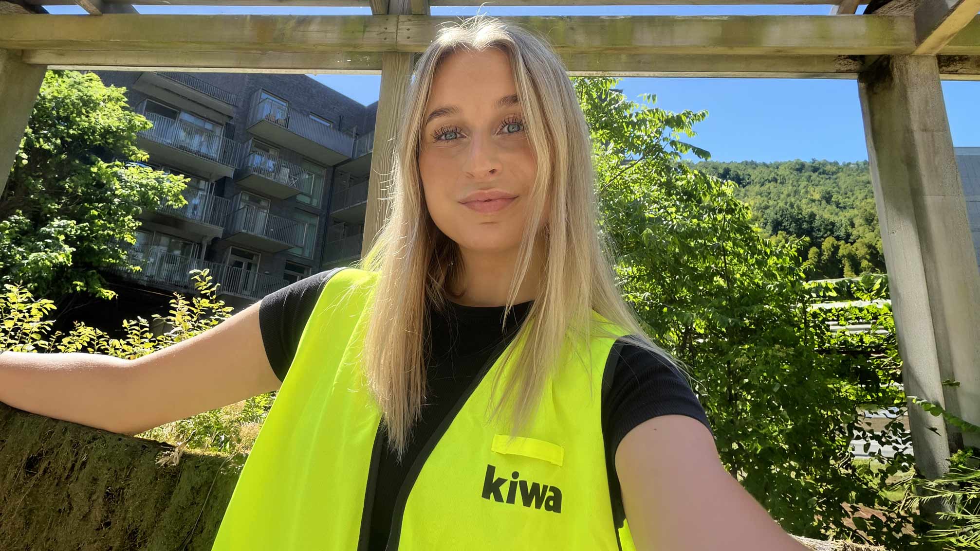 Porträtt av en person med blont hår och neongul Kiwa-väst utomhus i solljus.