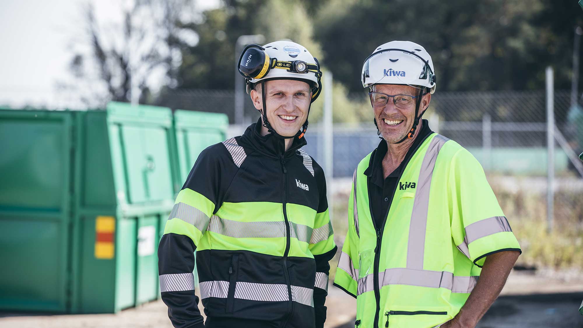Två Kiwakollegor i skyddskläder och hjälmar står utomhus på en arbetsplats.