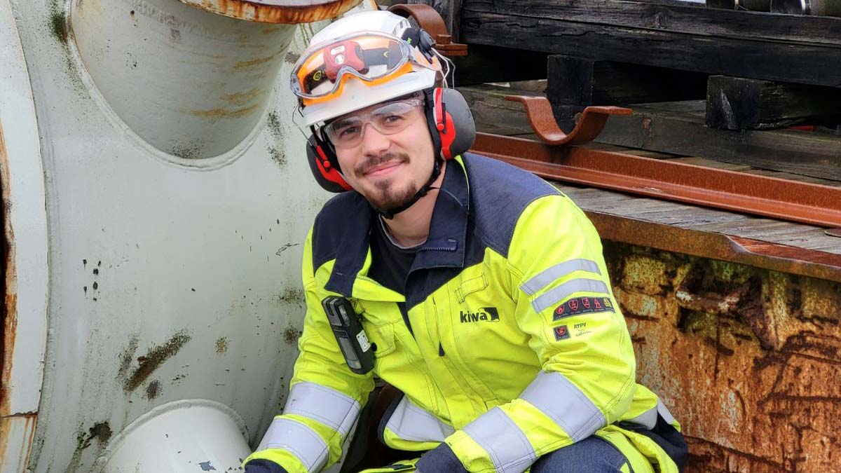 Person i varselkläder och hjälm vid en industriell miljö med rostiga metallkonstruktioner.