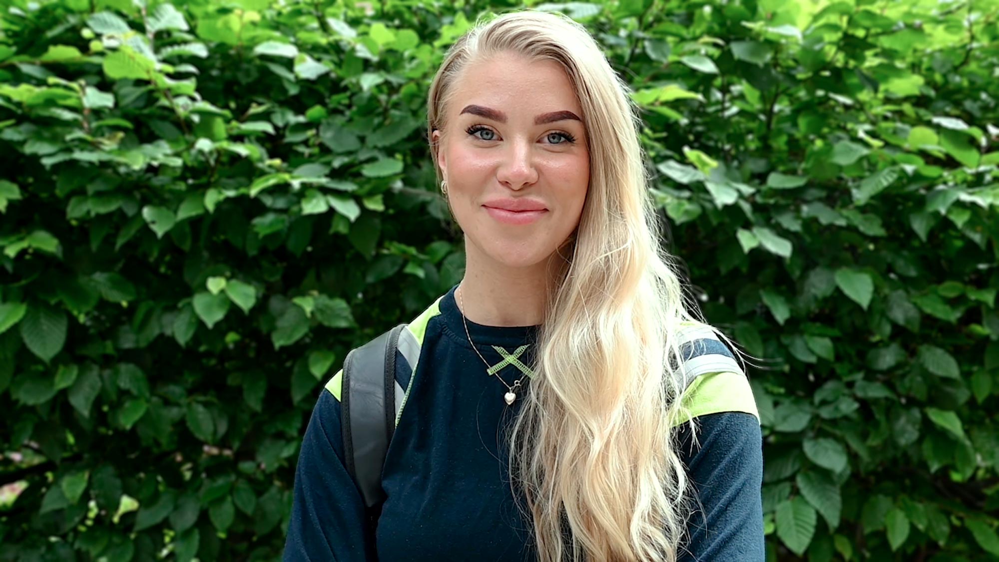 Porträtt av en kvinna i svart hoodie med neongula detaljer och logotypen Kiwa, stående inomhus.