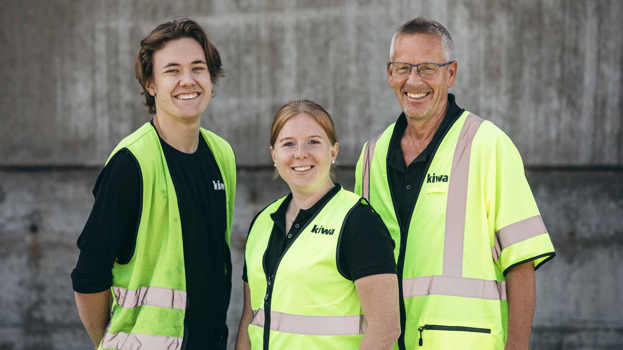 Tre personer i gula reflexvästar och svarta tröjor står framför en grå betongvägg.