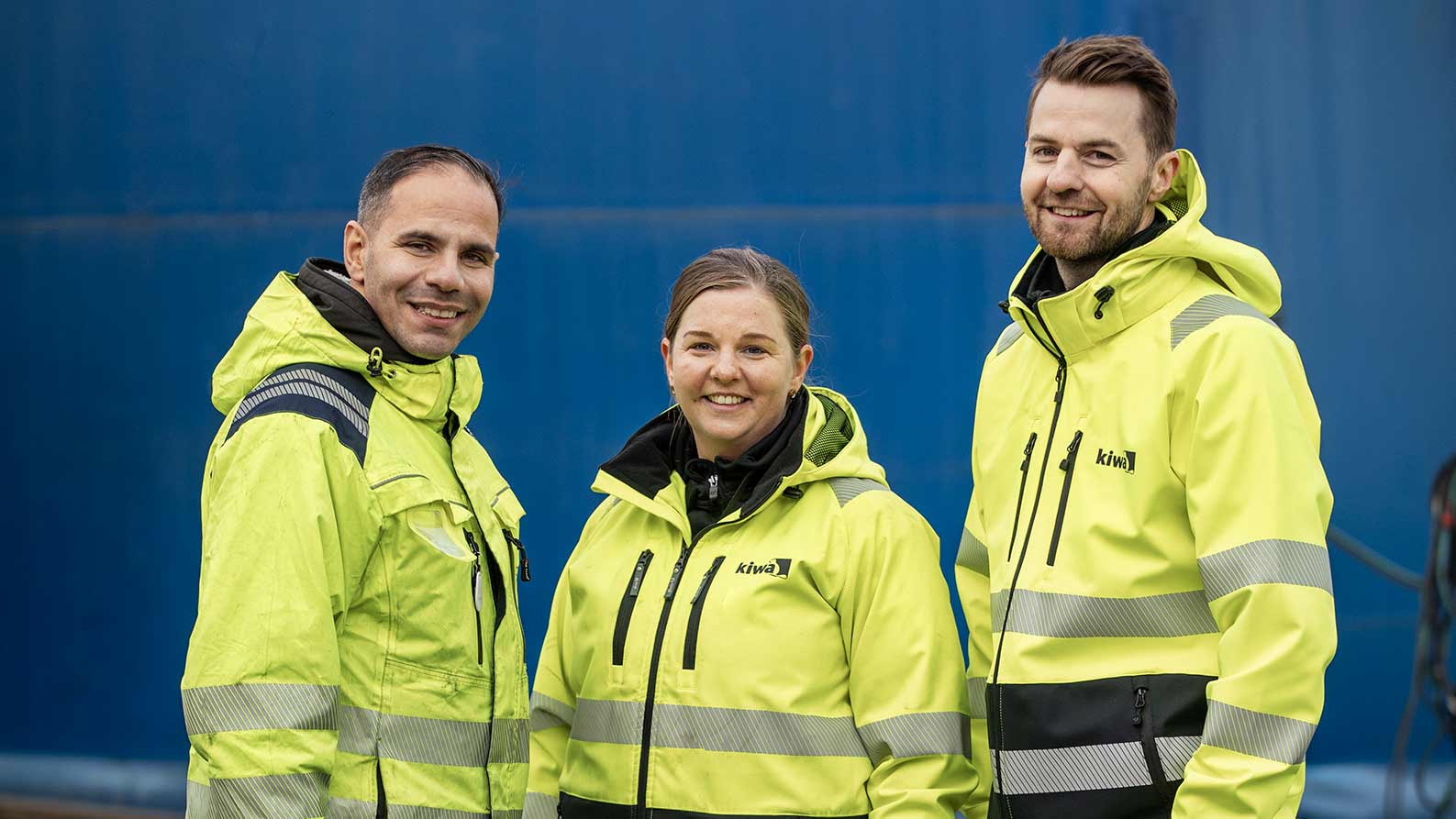 Tre Kiwakollegor i gula arbetsjackor står framför en blå bakgrund.