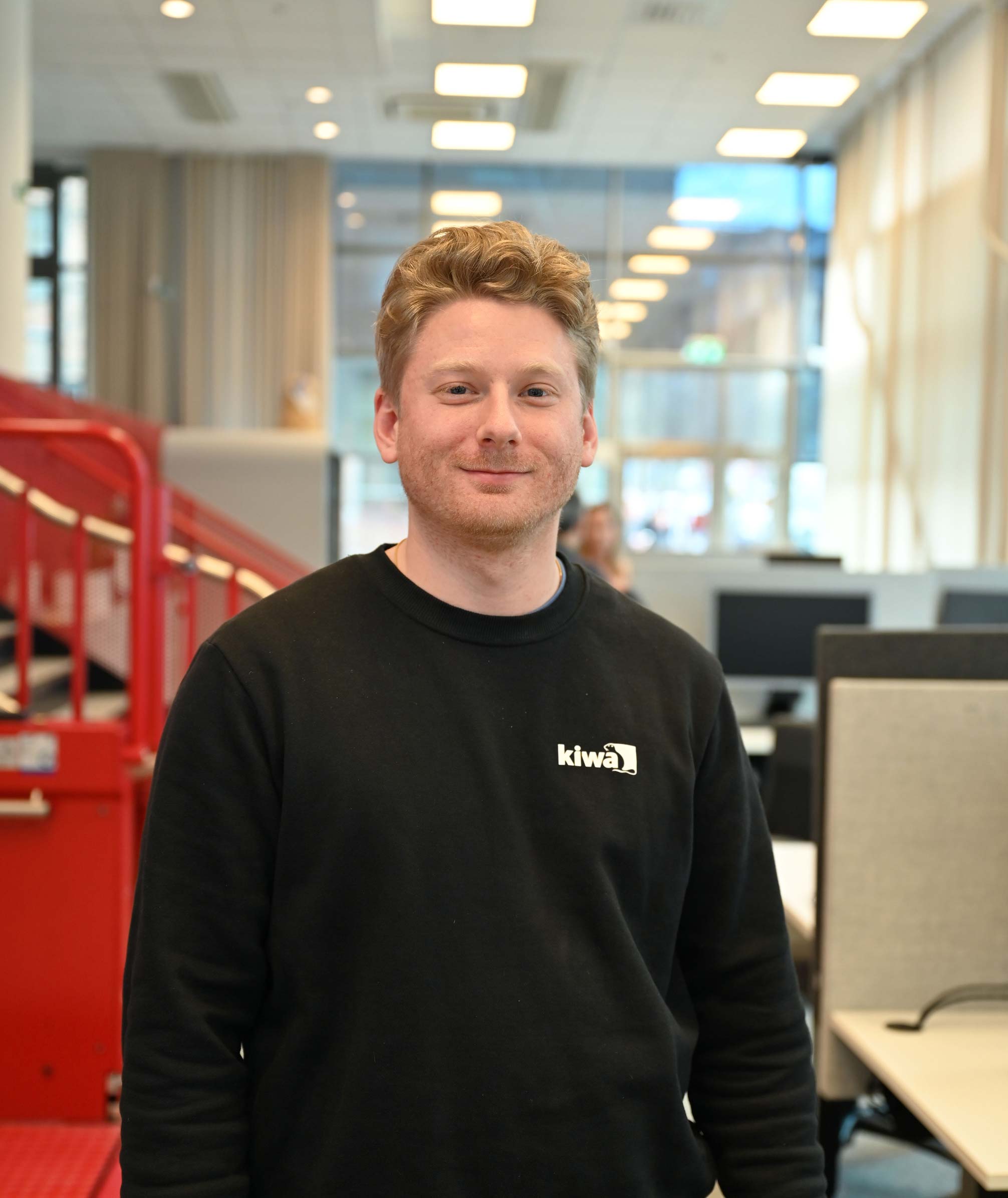 Porträtt av en person i svart tröja med logotyp, i ett kontor med datorarbetsplatser i bakgrunden.