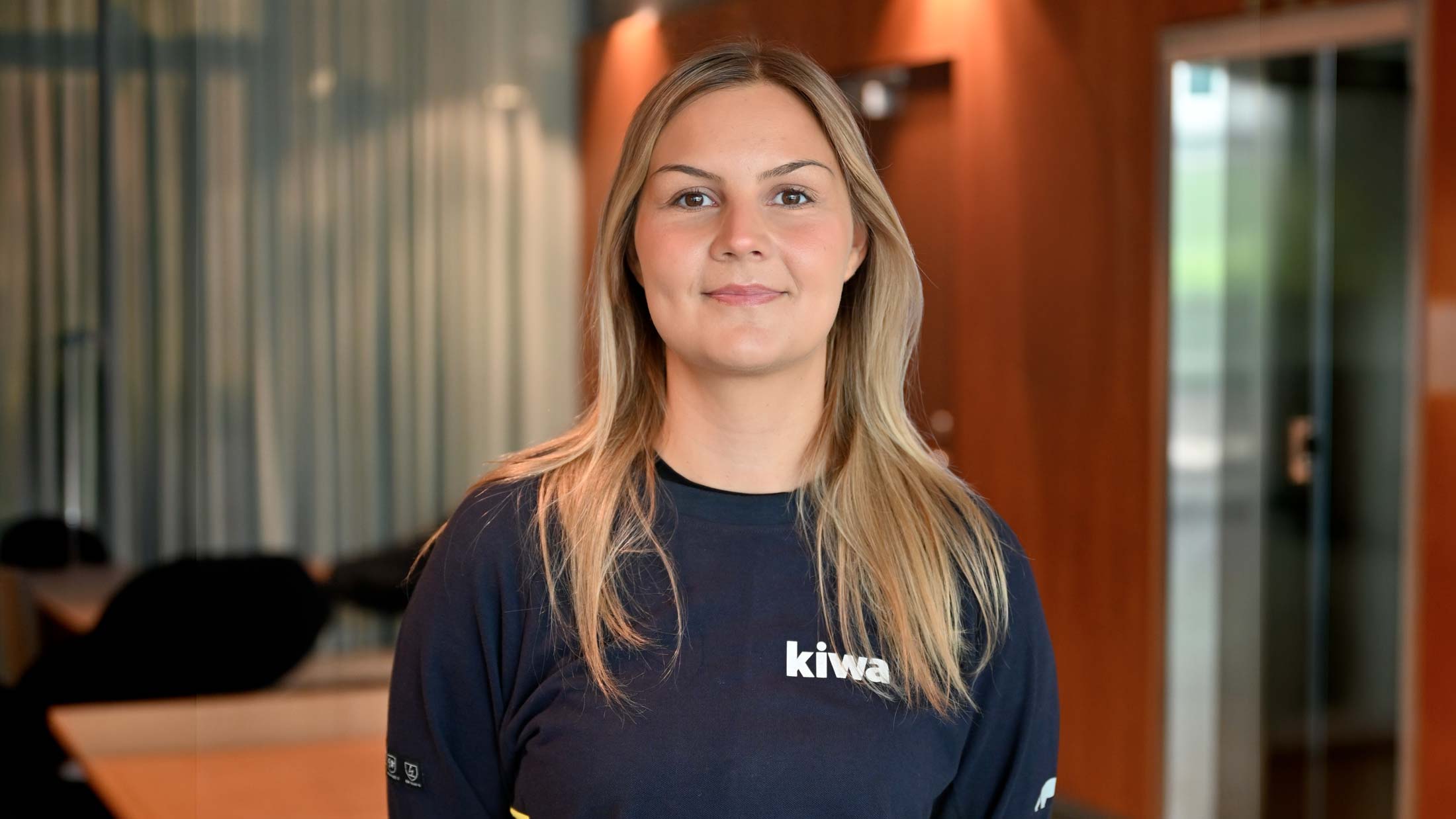 Porträtt av en kvinna med blont hår och mörk tröja med logotypen “kiwa”, stående inomhus i en modern miljö.