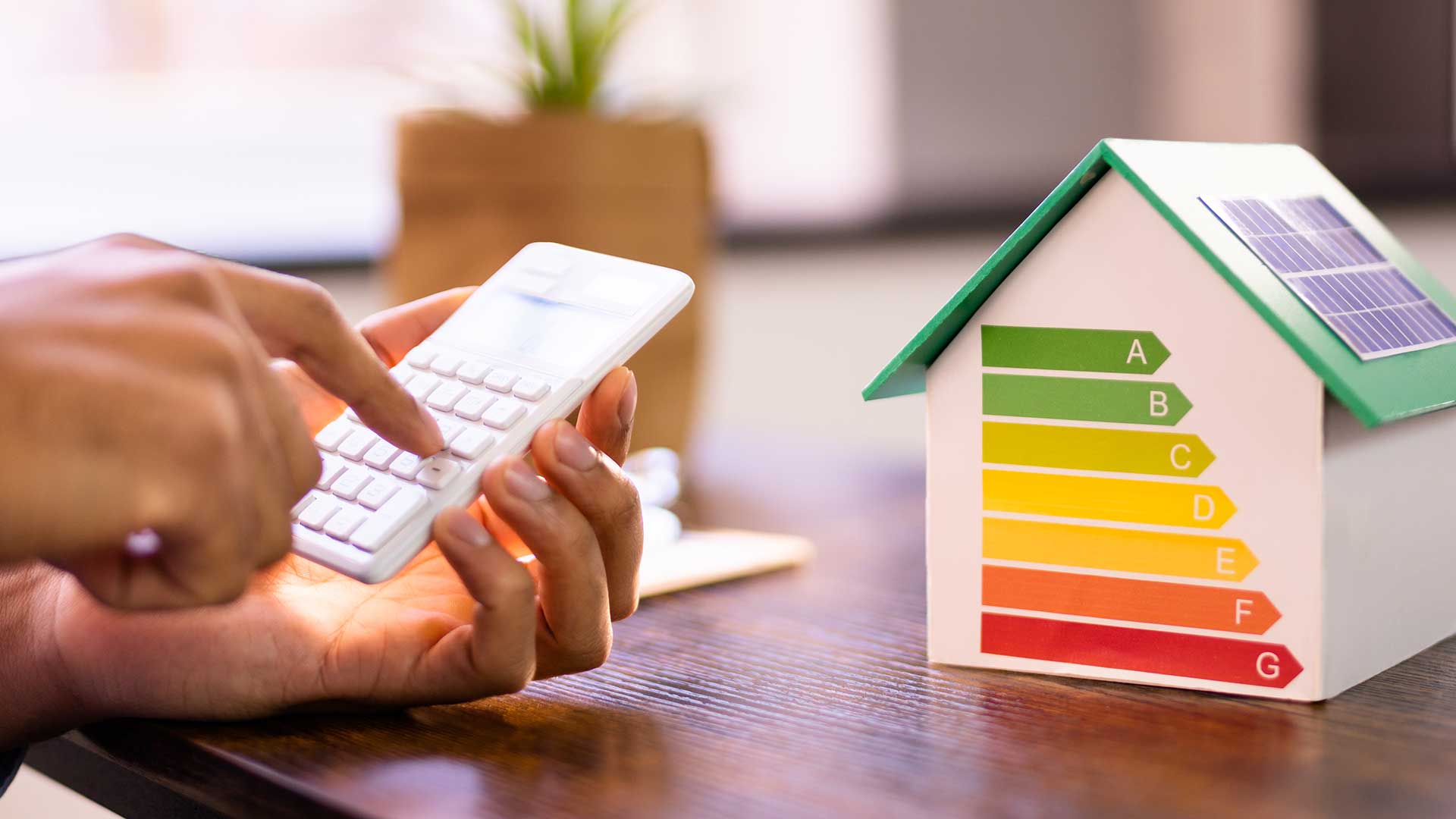Person som använder en miniräknare vid ett bord bredvid en modell av ett energieffektivt hus med solpaneler och energiklassning A–G.