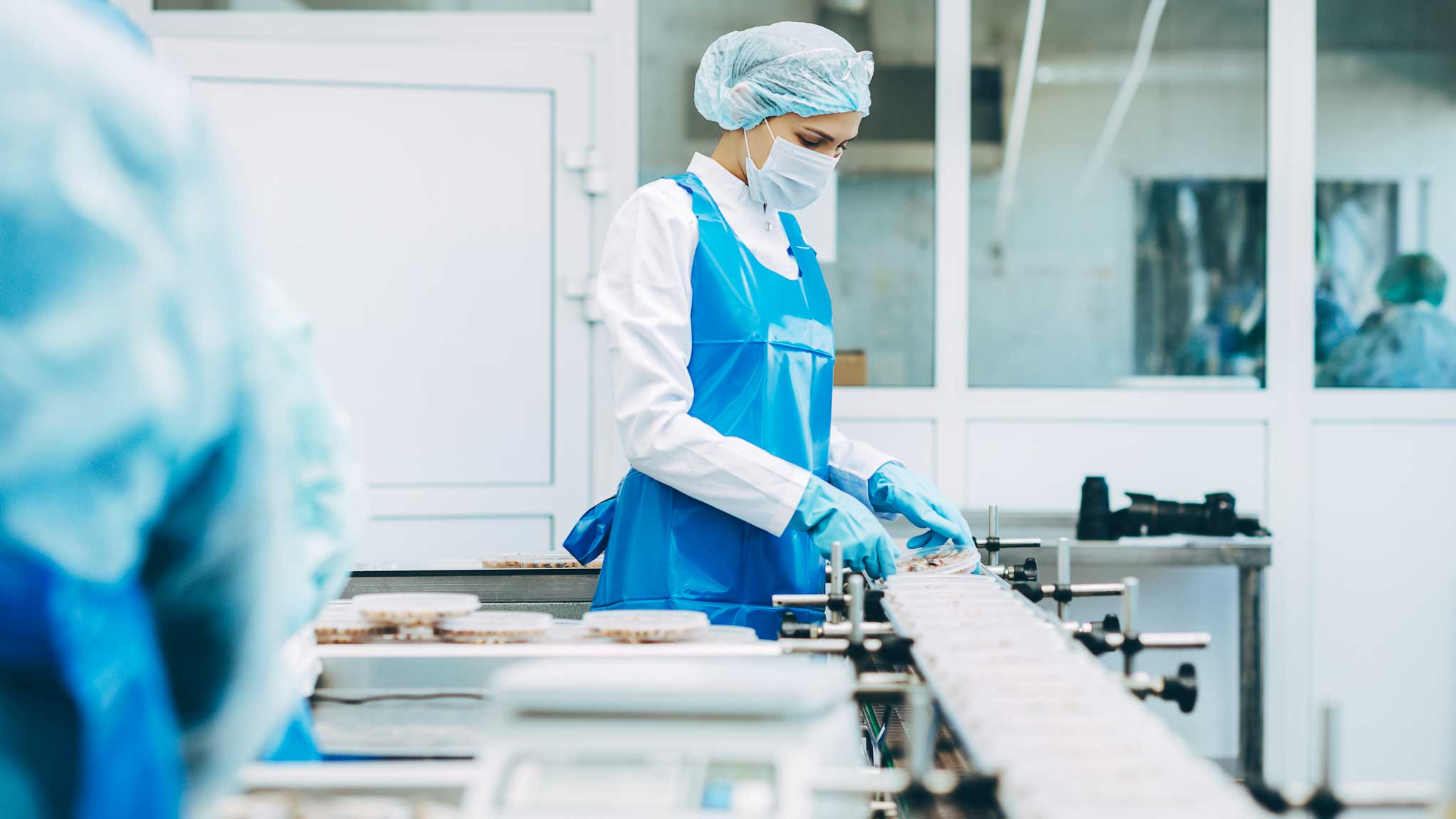 Kvinna i vit arbetsrock och hårnät kontrollerar matförpackningar i en produktionsmiljö, där bilden förmedlar vikten av livsmedelssäkerhet. 