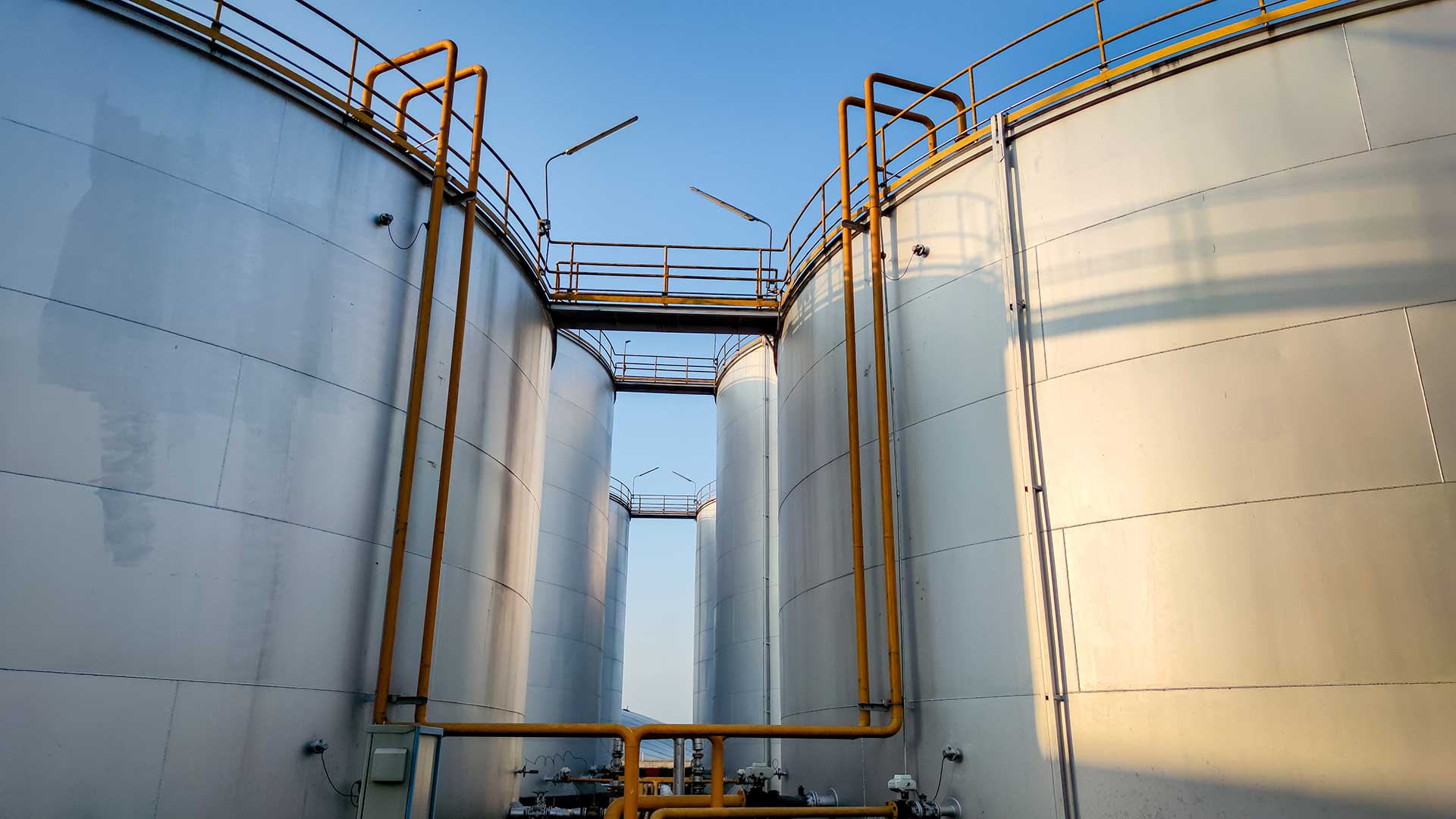 Stora industriella lagringstankar i metall med rör och gångbroar mot blå himmel.