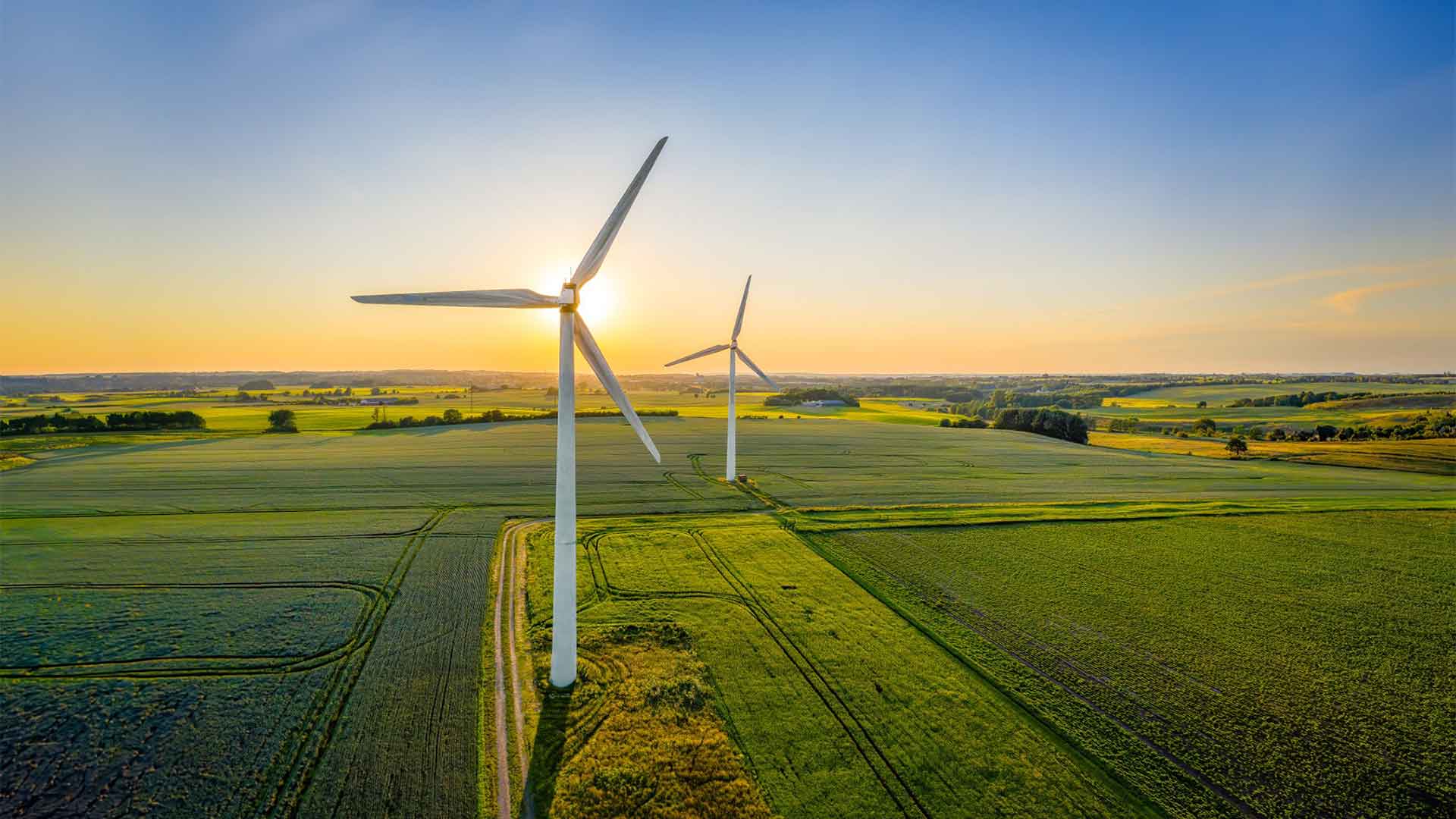 Vindkraftverk på åkermark i solnedgång med vidsträckt landskap och klar himmel i en bild som förmedlar behovet av att förlänga livslängden för befintliga vindkraftverk. 