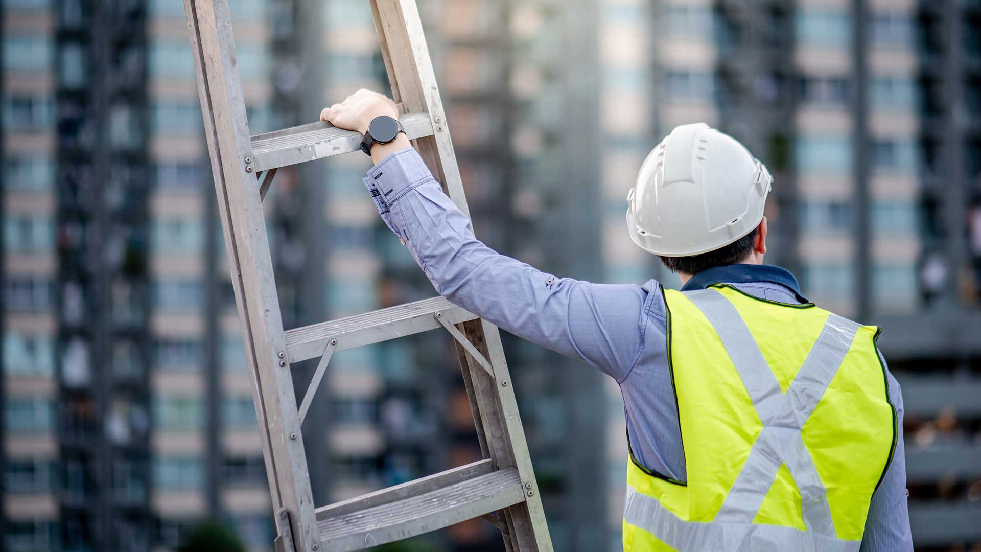 En man i skyddshjälm och varselväst håller i en stege och tittar bort mot bostadshus. 