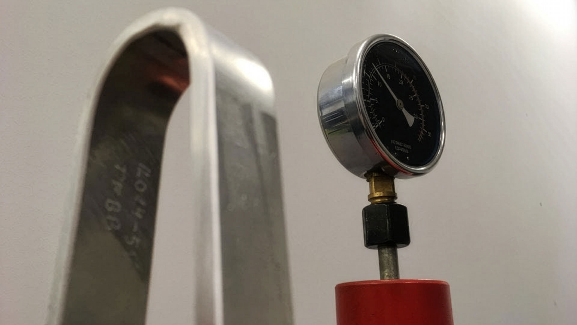 Manometer på en röd bas bredvid en böjd metallbåge med ingraverade siffror, som används för bockprovning.