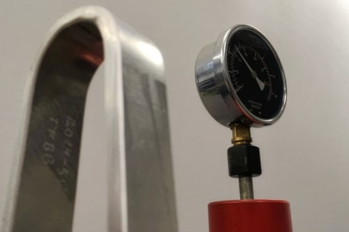 Manometer på en röd bas bredvid en böjd metallbåge med ingraverade siffror, som används för bockprovning.