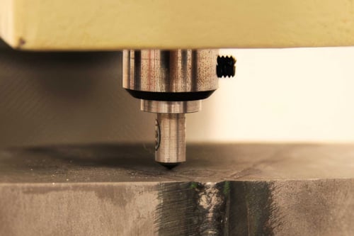 Närbild av en maskin som utför hårdhetsmätning där ett verktyg pressas mot en metallbit.