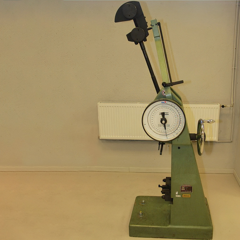 Grön slagprovningsutrustning, med mätare och arm, placerad på ett beige golv framför en ljusgrå vägg.