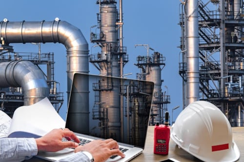 Ingenjör arbetar med dator med skyddshjälm bredvid, gas- och oljeraffinaderi visas i bakgrunden.