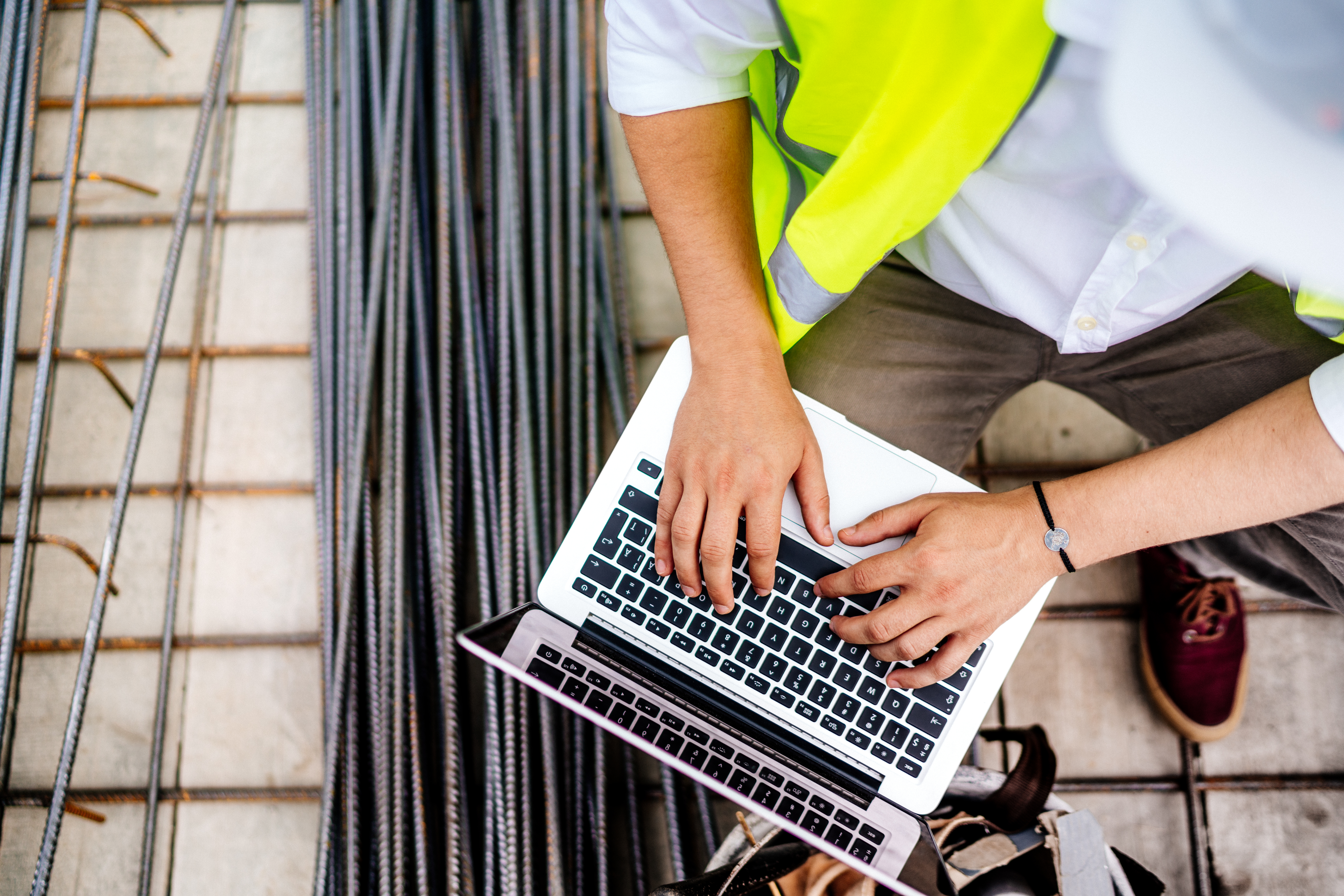Construction worker in a safety vest typing on a laptop beside stacked steel rebar at a building site