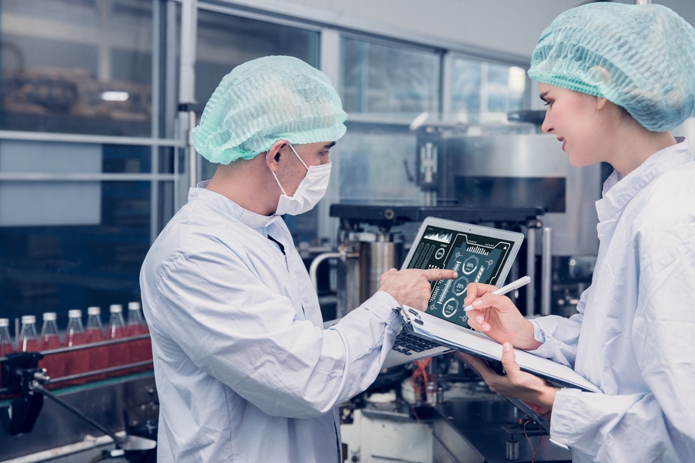 Two Kiwa ASI employees in protective clothing and hairnets in a manufacturing facility discuss data displayed on a tablet, with machinery and bottles visible in the background.