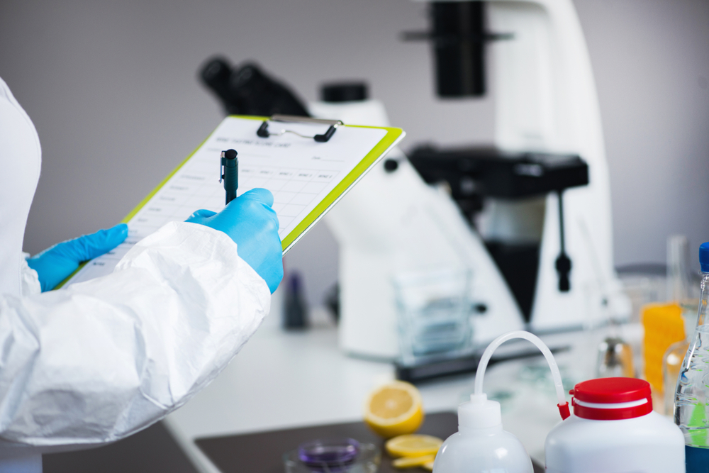 Persona con bata protectora y guantes azules escribiendo en un portapapeles en un laboratorio con un microscopio y materiales científicos en el fondo.