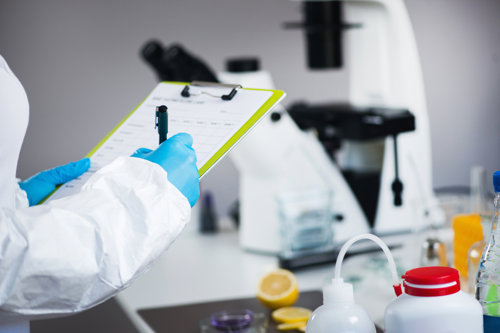 Persona con bata protectora y guantes azules escribiendo en un portapapeles en un laboratorio con un microscopio y materiales científicos en el fondo.