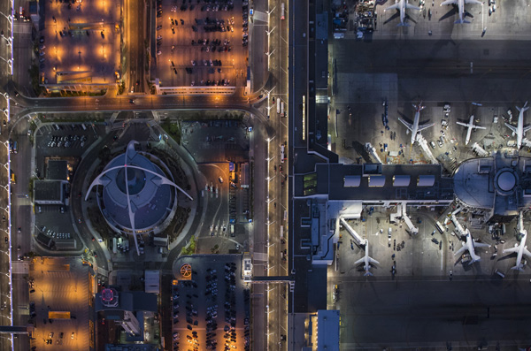 Aerial view of a busy airport terminal of LAX at night, with multiple airplanes parked at the gates and a brightly lit central structure surrounded by parking areas and roads.