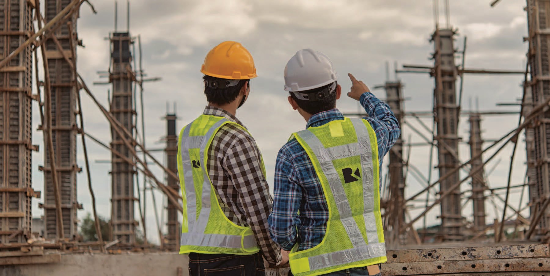 Two construction workers wearing hard hats and reflective safety vests stand at a building site, with one pointing toward scaffolding in the background.