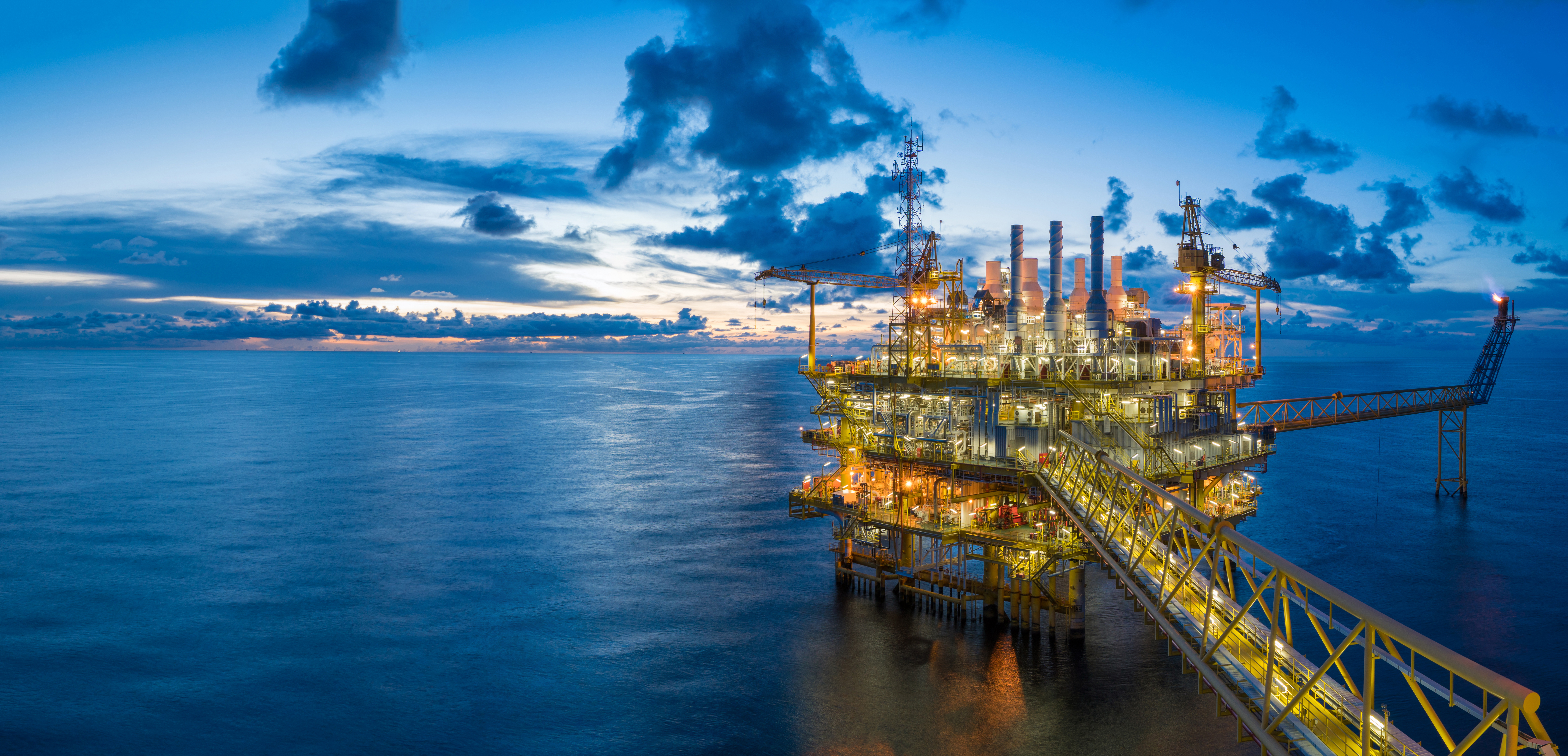 Offshore oil rig illuminated at dusk with a dramatic ocean backdrop and cloudy sky, showcasing industrial energy infrastructure in a marine environment.